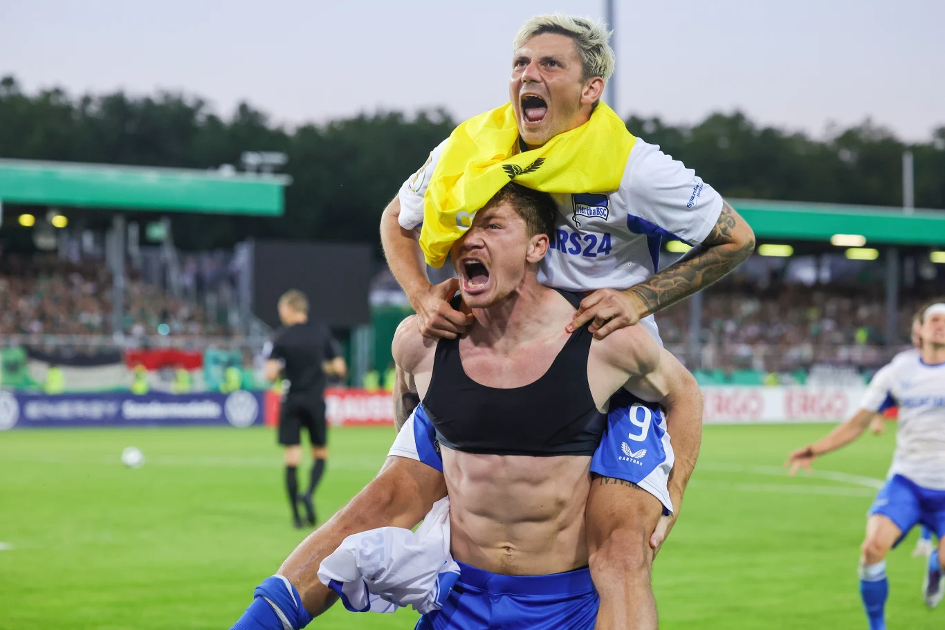 Dawid Kownacki (u góry) został bohaterem meczu z Holstein Kiel Dwóch piłkarzy drużyny świętuje na boisku, jeden niesie drugiego na barkach, obaj krzyczą z radości, wokół nich znajduje się murawa i trybuny pełne kibiców.