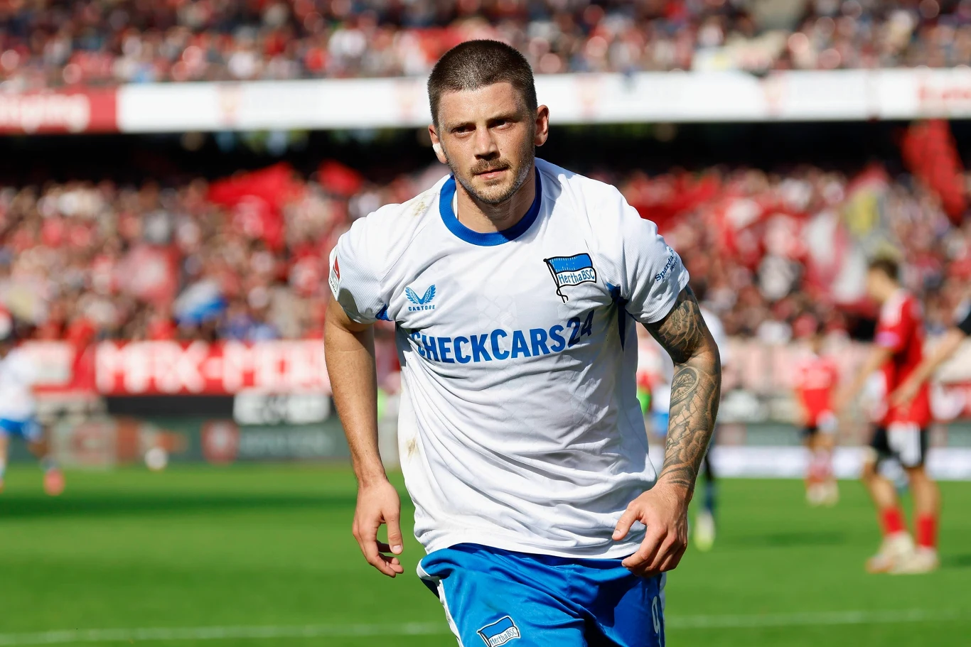 Dawid Kownacki Piłkarz w białej koszulce z niebieskimi elementami biegnie po boisku podczas meczu piłkarskiego, w tle rozmyta publiczność i inni zawodnicy.