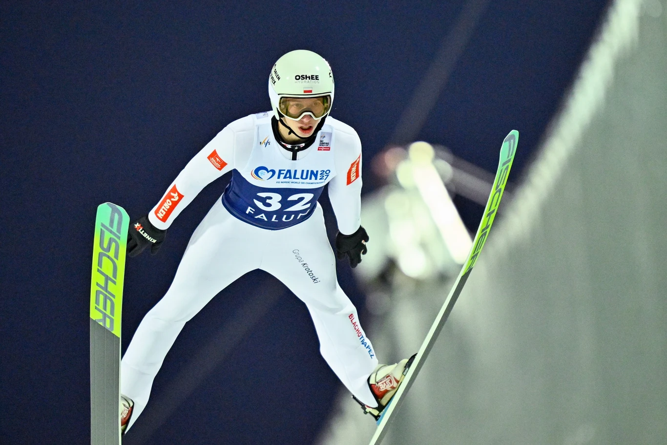 Kacper Tomasiak Skoczek narciarski w białym kombinezonie lecący w powietrzu podczas zawodów, trzymający narty równolegle do podłoża, w tle rozmazane światła i ciemne niebo.