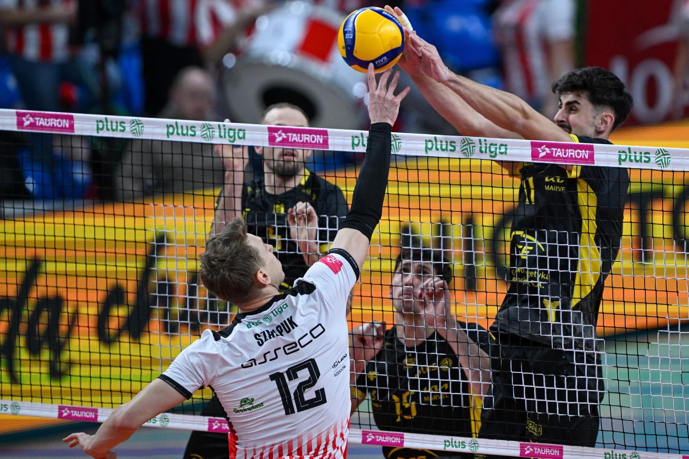 Artur Szalpuk kontra Antoine Pothron, mecz Asseco Resovia Rzeszów - PGE GiEK Skra Bełchatów Zawodnik w białej koszulce atakuje piłkę przy siatce, dwóch graczy w żółto-czarnych strojach próbuje zablokować akcję wyskakując do bloku po przeciwnej stronie siatki. W tle widoczna publiczność i reklamy.