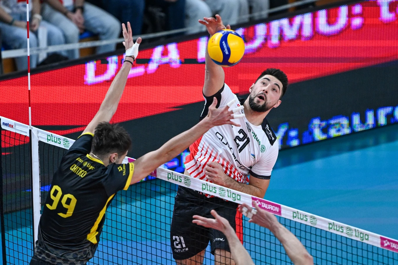 Atakuje Karol Butryn z Asseco Resovii Rzeszów Siatkarze rywalizują przy siatce podczas meczu, jeden zawodnik w białej koszulce z numerem 21 uderza piłkę ponad blokującego go przeciwnika w żółto-czarnej koszulce z numerem 99, widoczne ramiona innych graczy oraz fragment trybun.