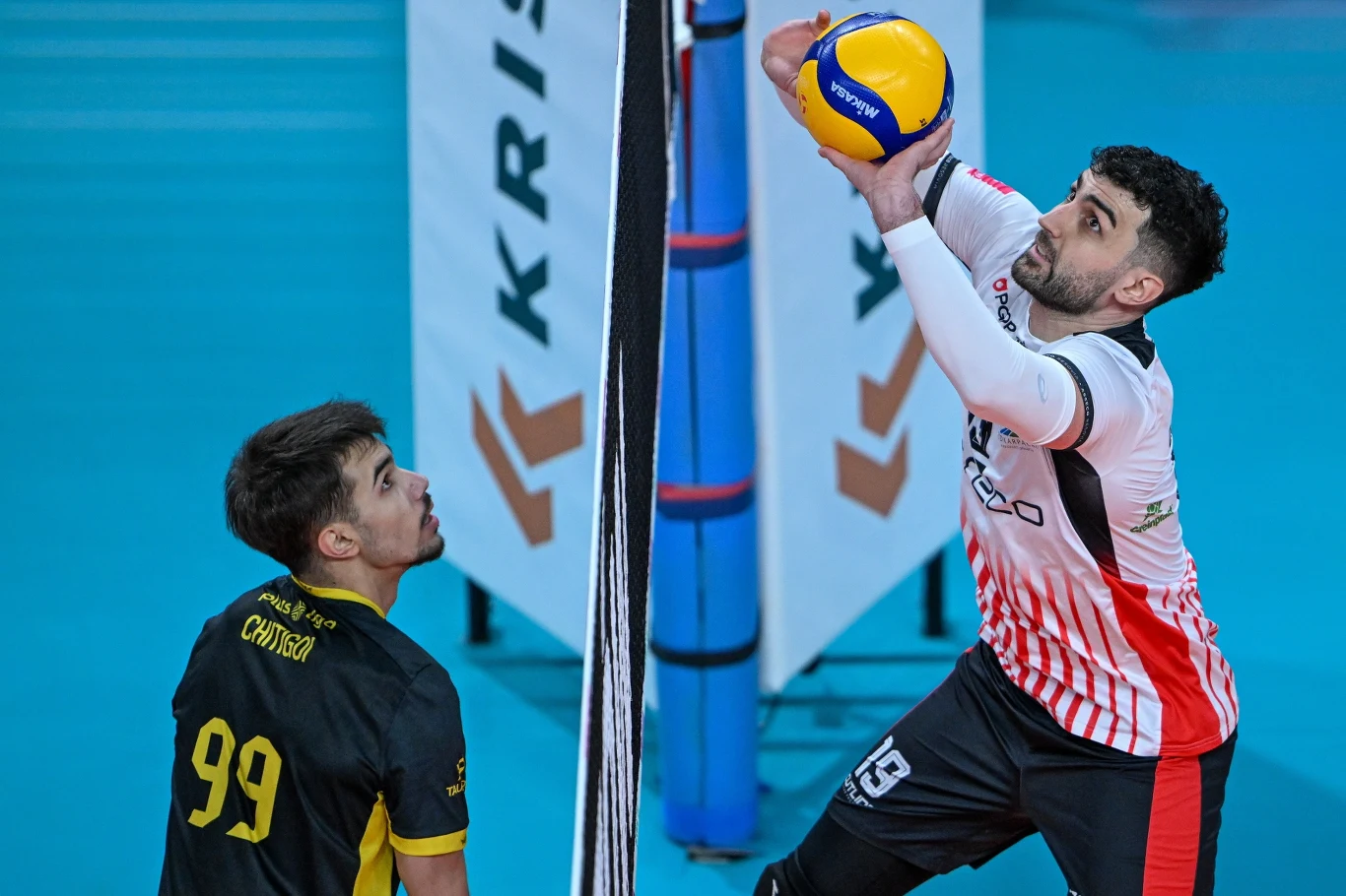 Daniel Chitigoi z PGE GiEK Skry Bełchatów (z lewej) i Marcin Janusz z Asseco Resovii Dwóch siatkarzy przy siatce podczas meczu, jeden z nich z czerwono-białej drużyny unosi się, przygotowując się do rozegrania piłki, zawodnik w czarno-żółtej koszulce z numerem 99 obserwuje akcję. Tło stanowi niebieska podłoga i sportowe banery.