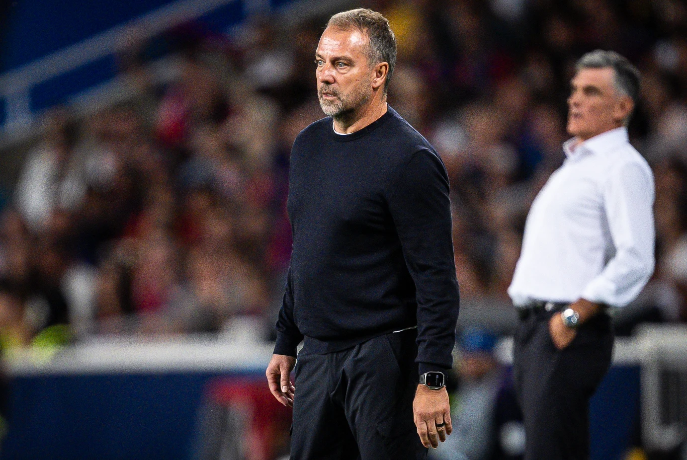 Hansi Flick Mężczyzna w ciemnym swetrze stoi na boisku, skupiony na obserwacji meczu piłkarskiego, w tle drugi mężczyzna patrzący w inną stronę, rozmyte trybuny pełne kibiców.