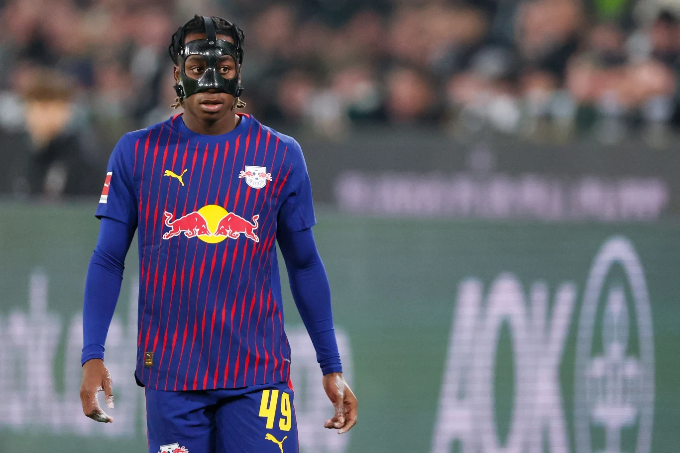 Yan Diomande Piłkarz drużyny RB Leipzig ubrany w sportowy strój z numerem 49, na twarzy czarna maska ochronna, w tle widownia stadionu sportowego.