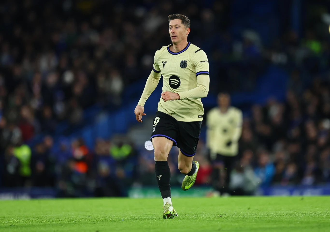 Robert Lewandowski Piłkarz w jasnej koszulce z numerem 9 biegnie po murawie podczas meczu piłkarskiego, w tle rozmazana publiczność stadionu.