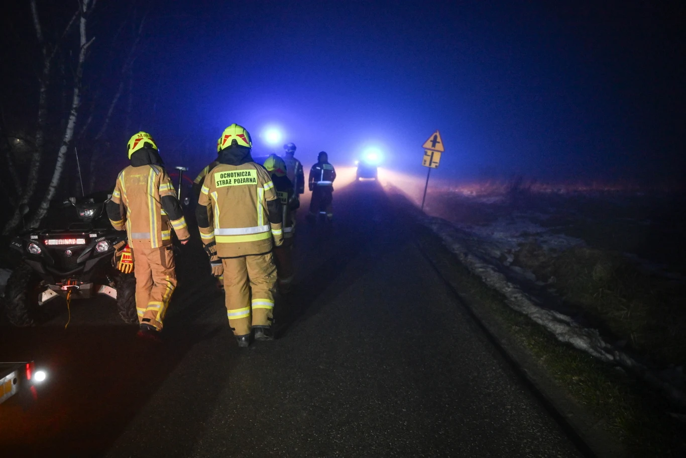 Strażacy w odblaskowych mundurach idą drogą w stronę samochodu ratowniczego z migającymi niebieskimi światłami, po bokach widoczny znak drogowy oraz zarośla, atmosfera wieczorna lub nocna z wyraźną mgłą.