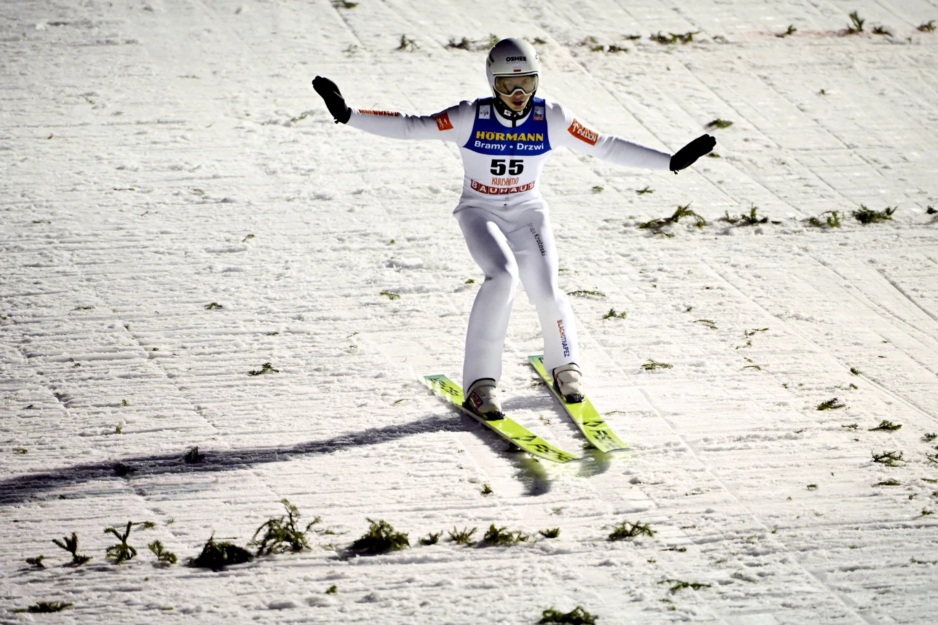 Skoczek narciarski ląduje na ośnieżonym zeskoku, unosząc ręce dla utrzymania równowagi, w tle widoczne ślady na śniegu oraz fragmenty zielonych gałązek ozdabiających zeskok.