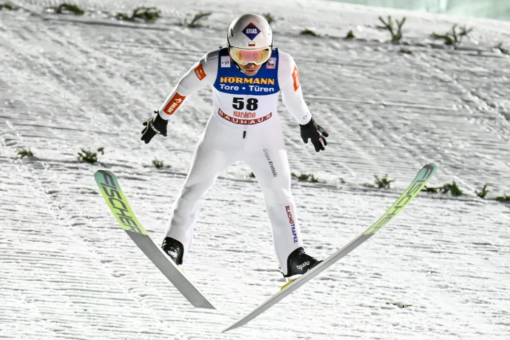 Kamil Stoch Skoczek narciarski w białym kombinezonie w locie nad skocznią, z numerem 58 na piersi, ujęcie wykonane podczas zawodów sportowych na tle ośnieżonego zeskoku.