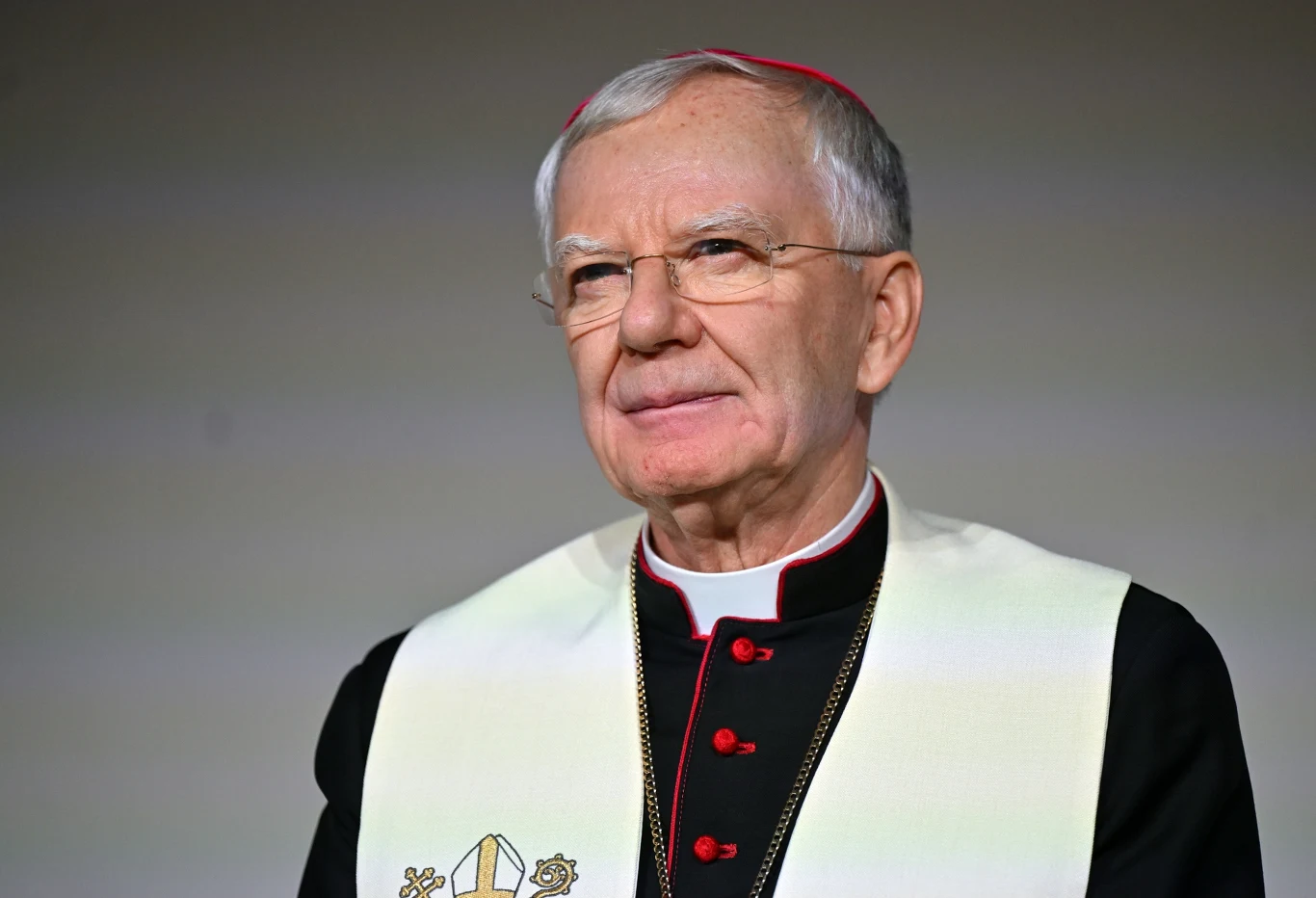 Abp Marek Jędraszewski wystosował list Starszy mężczyzna w stroju duchownego, uśmiechający się delikatnie, ubrany w czarną sutannę z czerwonymi guzikami i białą stułą, na jednolitym, szarym tle.