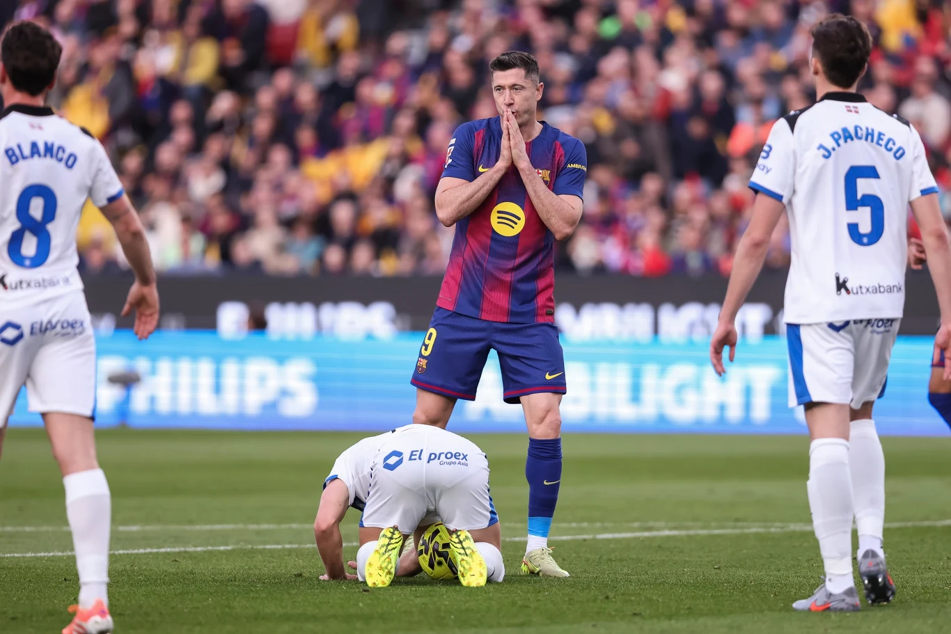 Robert Lewandowski podczas meczu z Deportivo Alaves Piłkarz w stroju FC Barcelona w centrum boiska wyraża emocje, stojąc z połączonymi dłońmi przy ustach. Obok niego zawodnik drużyny przeciwnej w białym stroju klęczy pochylony do przodu, a na drugim planie widoczni są kolejni piłkarze i tłum kibiców na ...