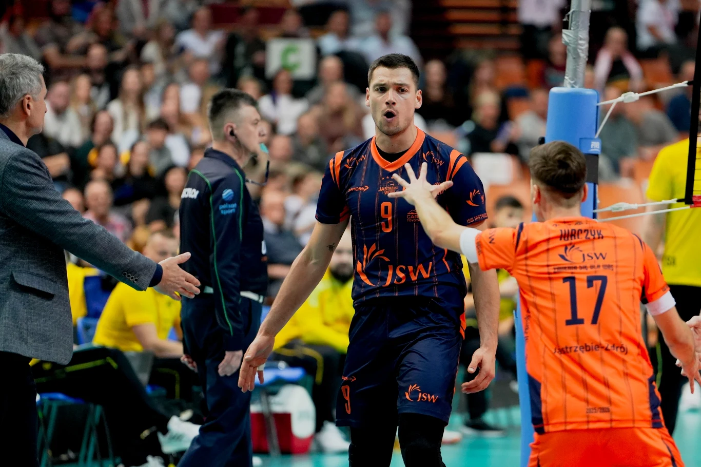 Zawodnik drużyny siatkarskiej w granatowo-pomarańczowym stroju wykazuje emocje na boisku po udanej akcji, wokół niego pozostali gracze oraz sędziowie, w tle publiczność obserwuje przebieg meczu.