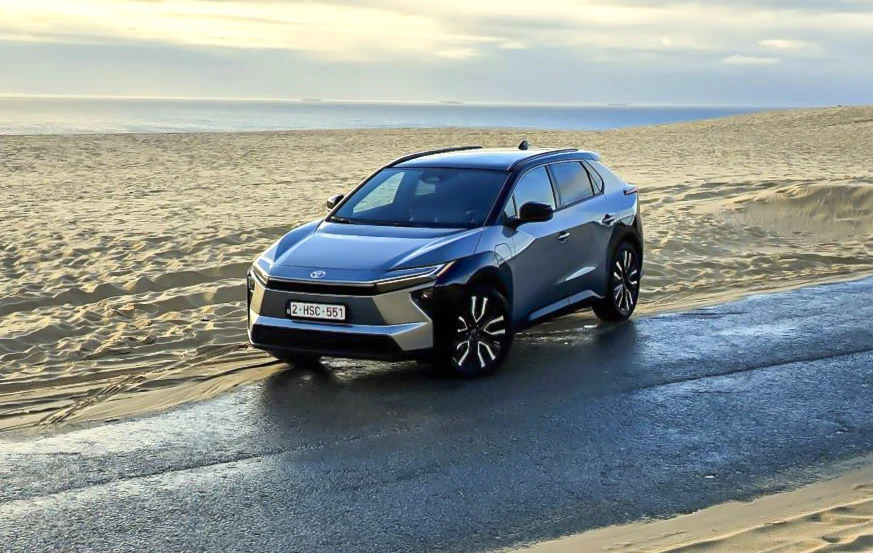 Nowoczesny samochód SUV zaparkowany na mokrej drodze biegnącej przez pustynną plażę przy spokojnym morzu, pod pochmurnym niebem.