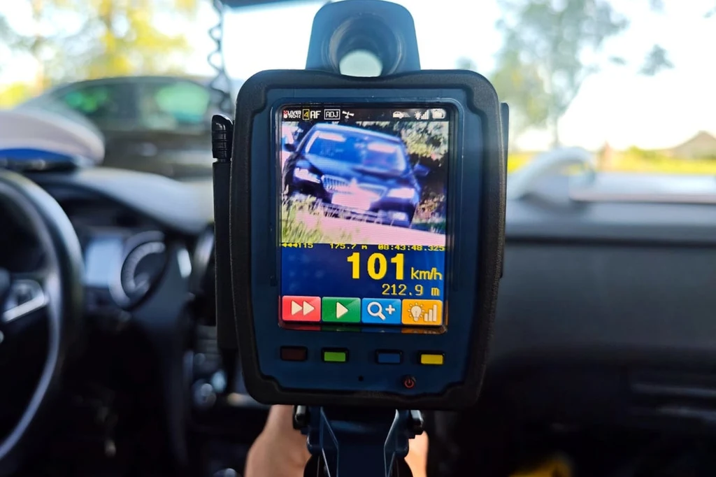 Ręczny miernik prędkości rejestrujący samochód osobowy jadący z prędkością 101 km/h, wyświetlacz pokazuje odległość do pojazdu oraz dodatkowe informacje wizualne, wnętrze radiowozu w tle.