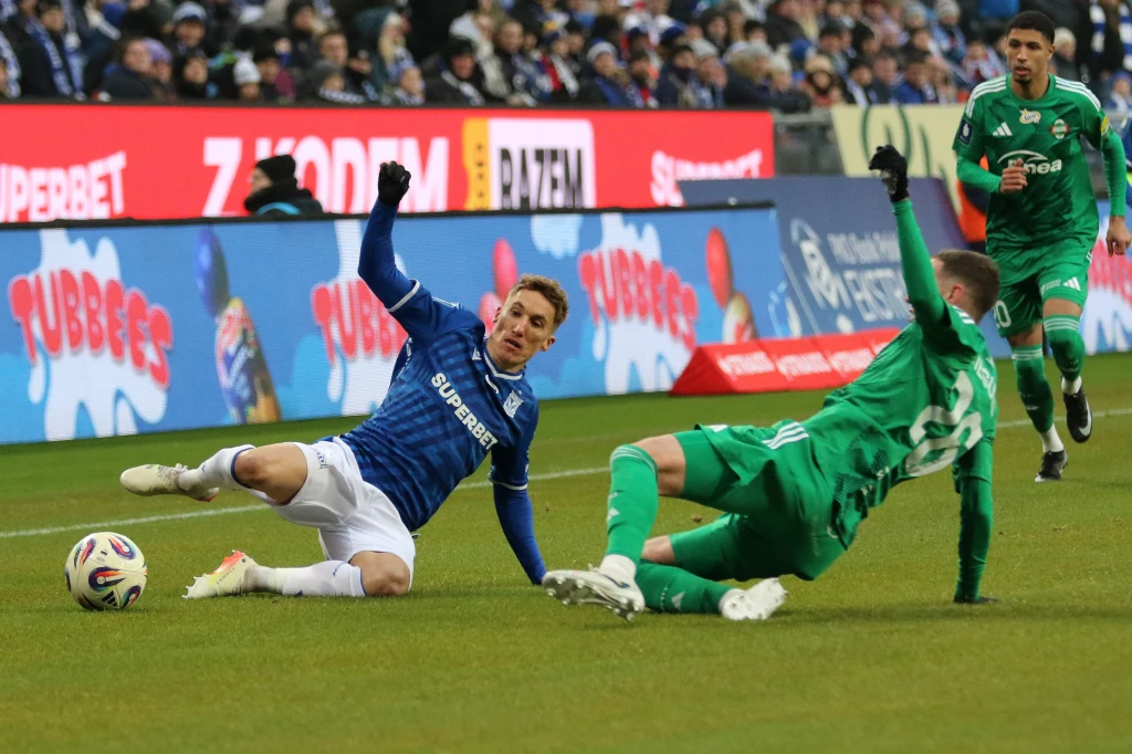 Dwóch piłkarzy walczy o piłkę podczas meczu, jeden ubrany w niebieski strój leży na murawie, drugi w zielonym wykonuje wślizg, w tle widoczni pozostali zawodnicy oraz kibice na trybunach.