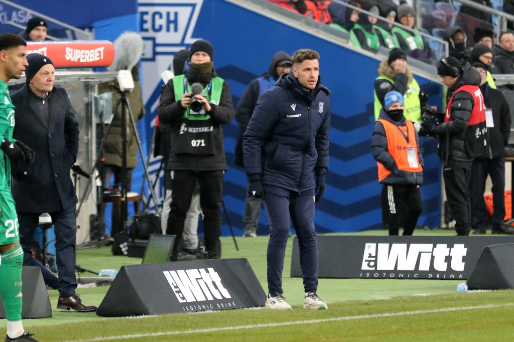 Trener piłkarski w sportowym stroju gestykuluje przy linii bocznej boiska podczas meczu, w tle widoczni są zawodnicy, sędziowie techniczni oraz obsługa techniczna stadionu, a także tablice reklamowe.