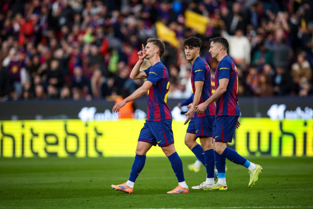 Piłkarze w koszulkach FC Barcelony świętują zdobycie gola na stadionie, jeden z zawodników wykonuje gest dłonią przy ustach, a za nimi widać tłum kibiców.