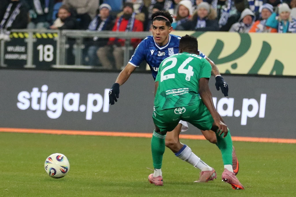 Dwóch piłkarzy w akcji na murawie podczas meczu piłkarskiego, jeden zawodnik w niebieskim stroju, drugi w zielonym, skupieni na piłce, za nimi widoczni kibice na trybunach.