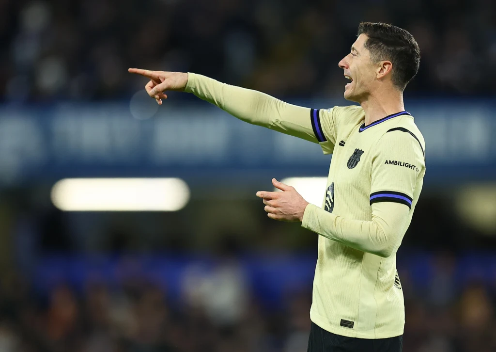 Piłkarz w jasnożółtej koszulce w trakcie meczu wskazuje coś ręką, otwierając usta, prawdopodobnie wydając polecenia lub komentując sytuację boiskową, w tle widoczna rozmyta publiczność.