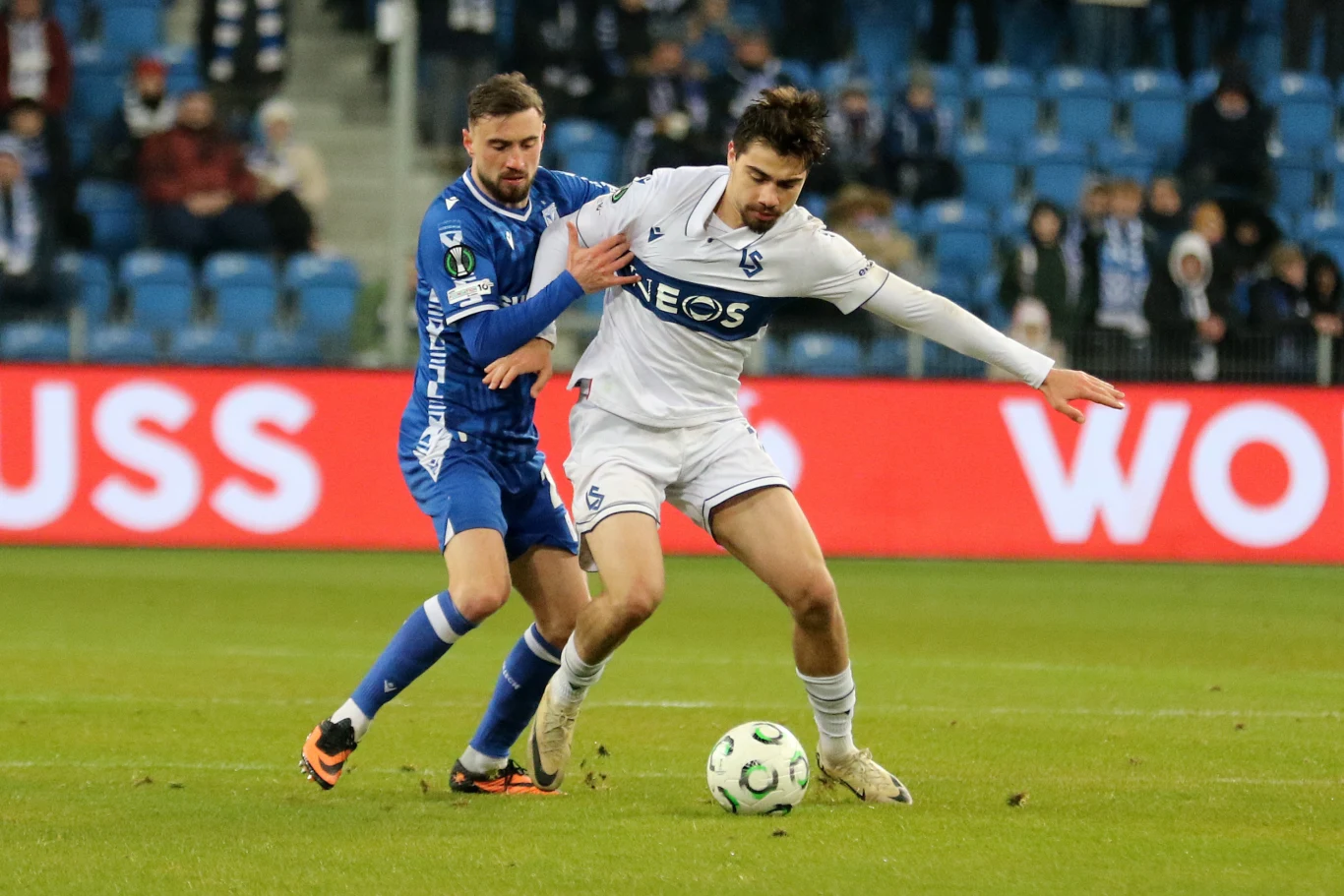 Dwóch piłkarzy walczących o piłkę podczas meczu piłkarskiego, jeden ubrany w niebieski strój, drugi w biały, trybuny wypełnione kibicami w tle.