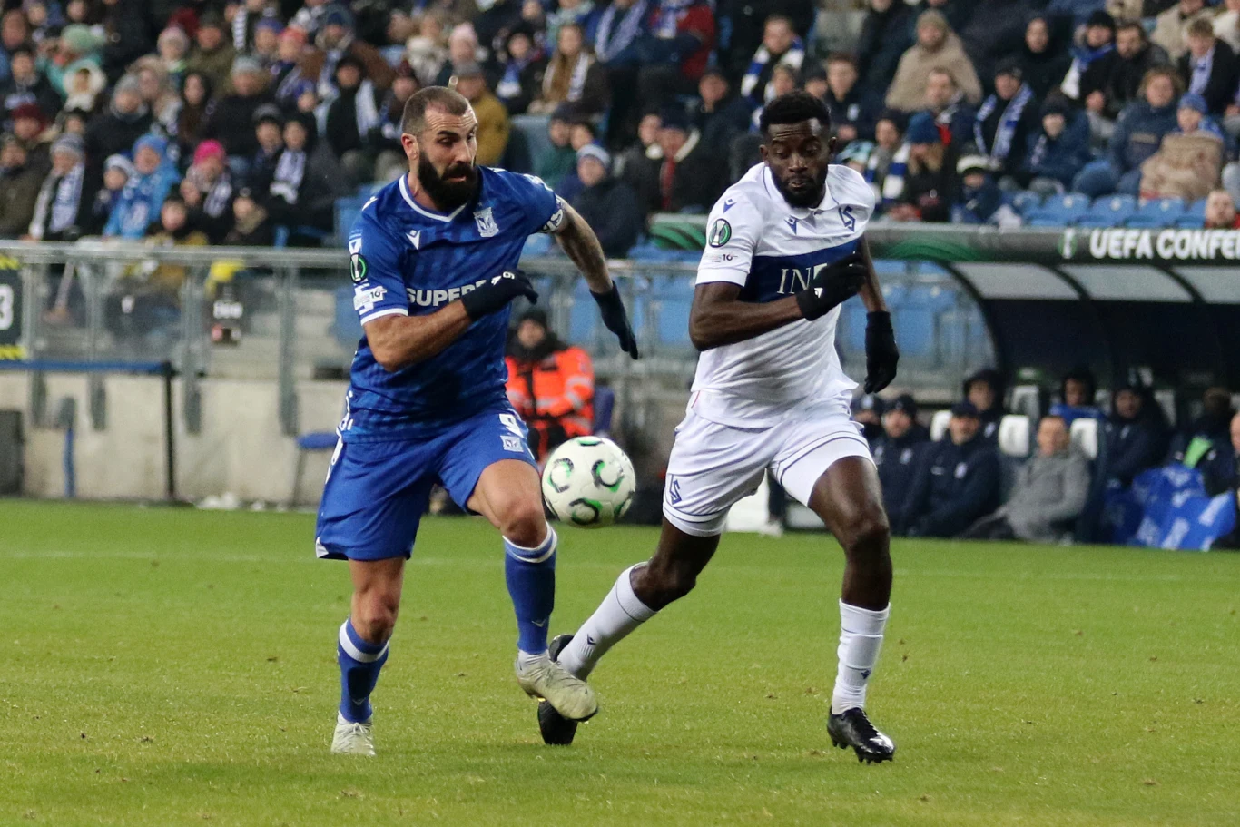 Dwóch piłkarzy rywalizuje o piłkę podczas meczu piłki nożnej na stadionie, wokół zasiada liczna publiczność, a w tle widoczne są banery z napisem UEFA Conference League.