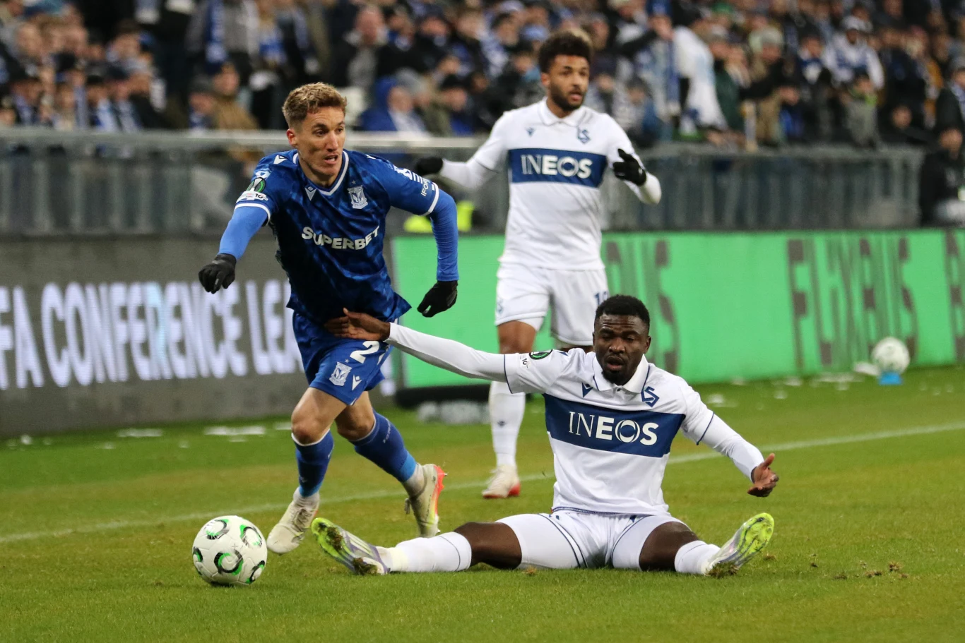 Zawodnik w niebieskim stroju próbuje wyminąć piłkarza w białym stroju siedzącego na murawie podczas meczu piłkarskiego, w tle widoczna publiczność na trybunach.