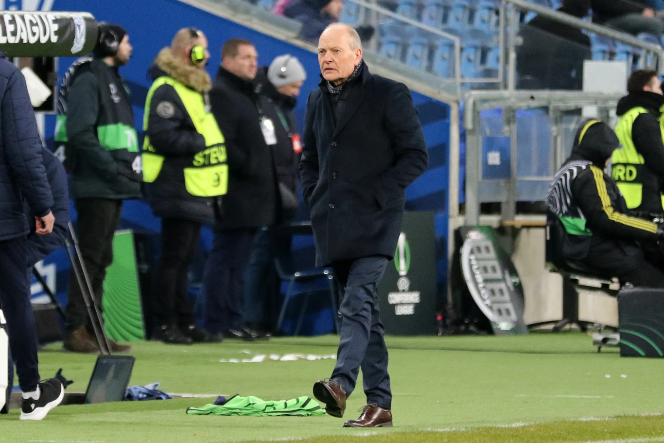 Trener drużyny piłkarskiej stoi przy linii bocznej boiska, ubrany w elegancki płaszcz, w tle widoczni są stewardzi w żółtych kamizelkach oraz kilka osób związanych z obsługą meczu.