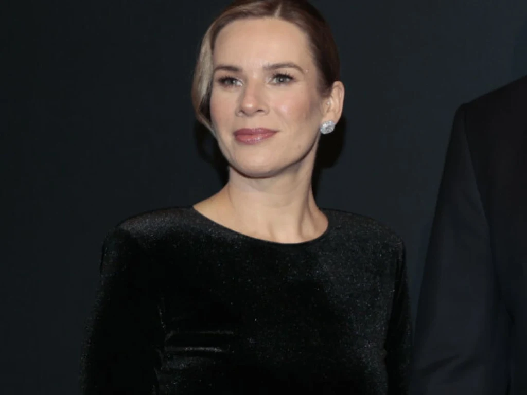 Kobieta w czarnej, eleganckiej sukni stoi na tle ciemnego tła, ma upięte włosy i delikatny makijaż, na uchu błyszcząca biżuteria.