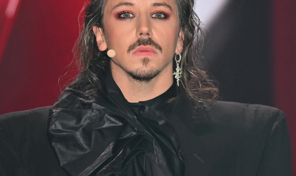 Osoba z długimi, ciemnymi włosami, wyrazistym makijażem z czerwonym cieniem na powiekach, ubrana w czarny strój z ozdobnym żabotem. Widoczna kolczyk w uchu oraz mikrofon przy twarzy. Tło w odcieniach czerwieni i czerni.