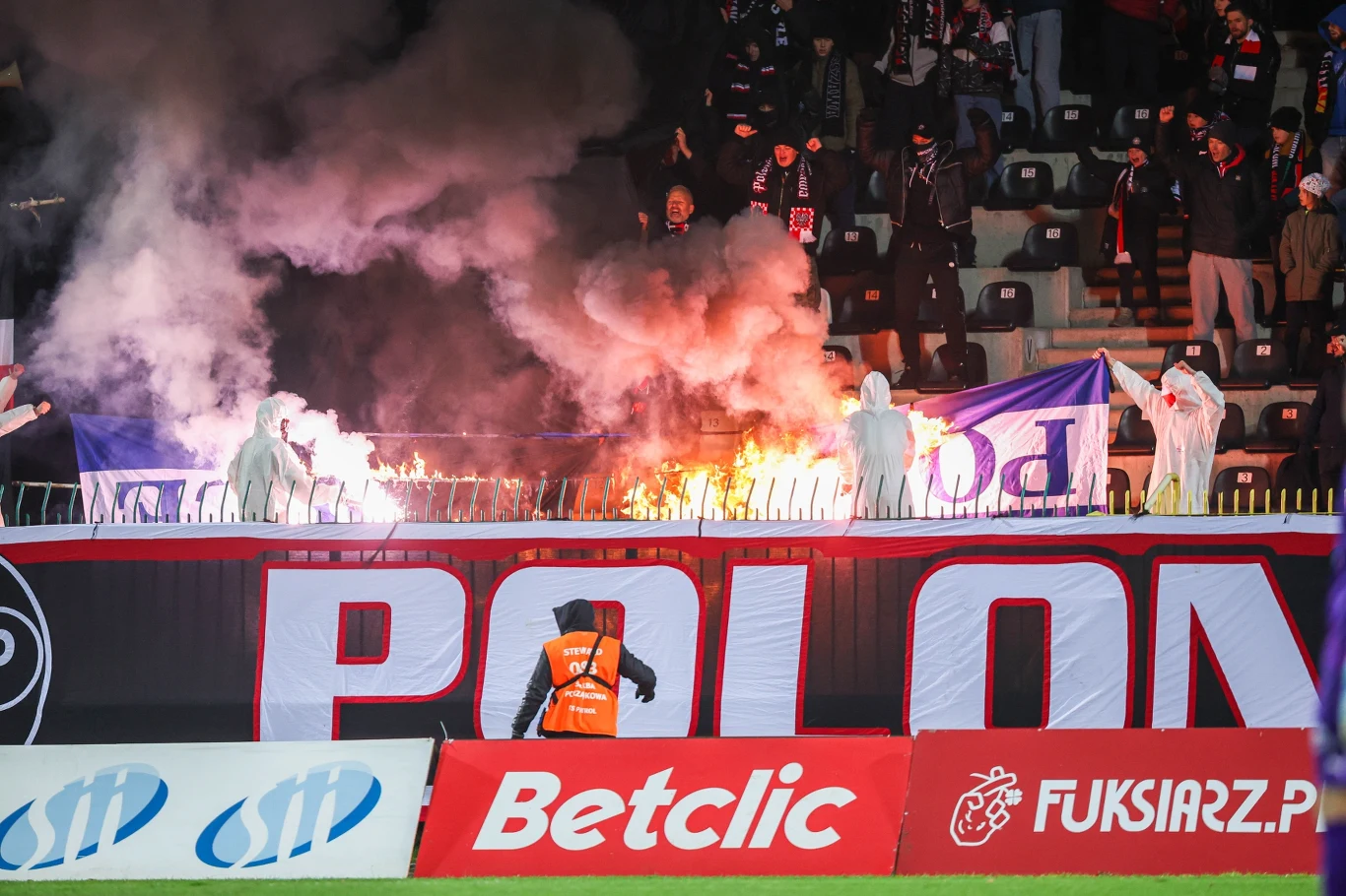 Grupa kibiców ubranych w białe kombinezony ochronne trzyma race świetlne na trybunach stadionu, unoszący się dym i ogień podkreślają emocje i atmosferę wydarzenia sportowego. Widoczne są barwy klubowe oraz banery reklamowe na pierwszym planie.