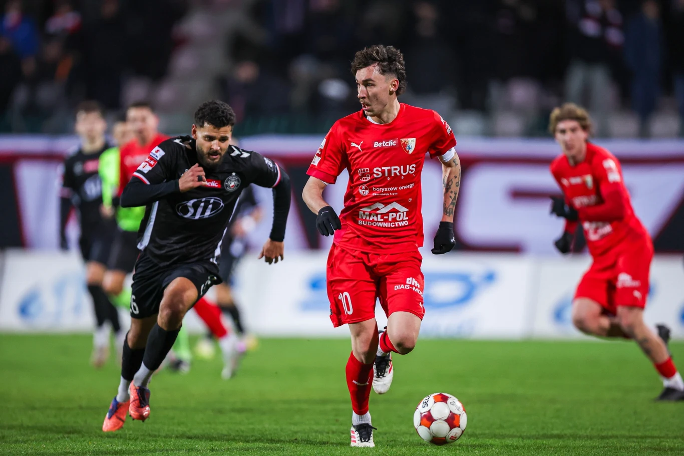 Dwóch piłkarzy w trakcie dynamicznego meczu piłki nożnej, jeden w czerwonym stroju prowadzi piłkę, a drugi w czarnym stroju próbuje go dogonić. W tle inni zawodnicy na boisku oraz rozmyta publiczność na trybunach.