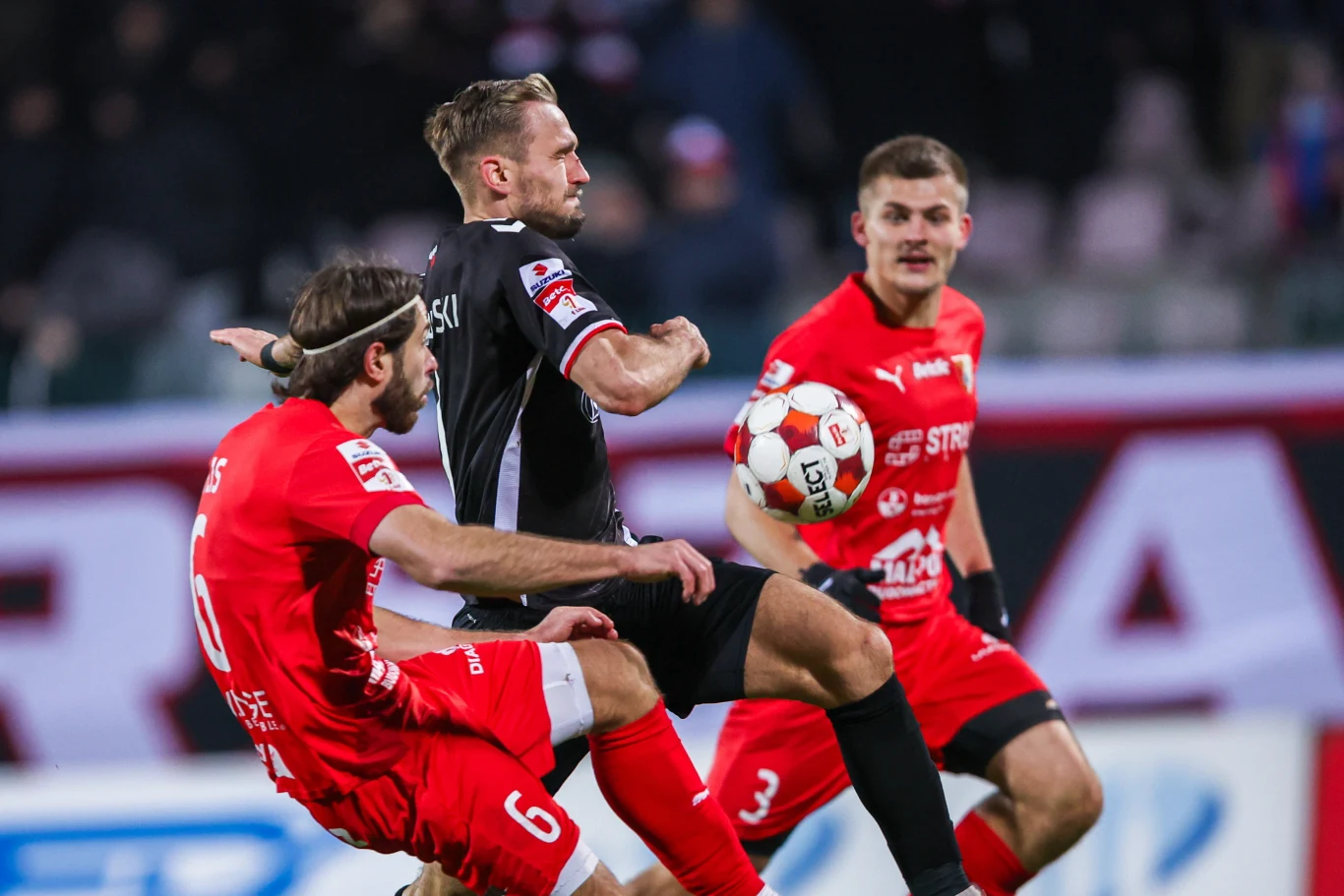 Zacięta akcja podczas meczu piłki nożnej, trzech zawodników walczy o piłkę przy intensywnym kontakcie. Dwóch piłkarzy w czerwonych strojach i jeden w czarnym są w centralnym punkcie kadru. Piłka znajduje się w powietrzu pomiędzy nimi.