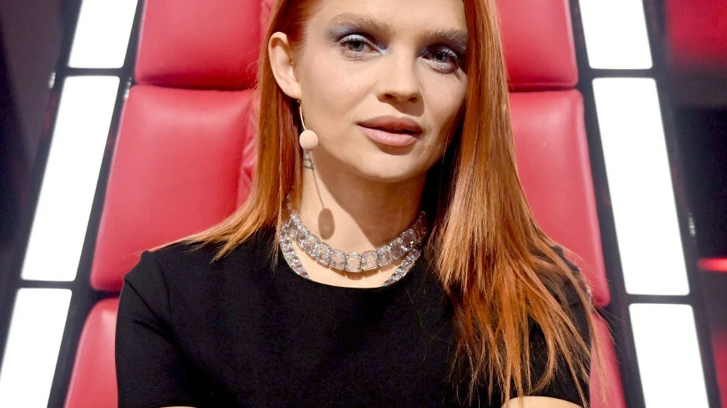 Margaret Kobieta z rudymi włosami w eleganckim makijażu siedzi na dużym czerwonym fotelu, ubrana w czarną bluzkę i srebrny naszyjnik, z mikrofonem przypiętym do ucha, tło stanowią podświetlone, pionowe panele.