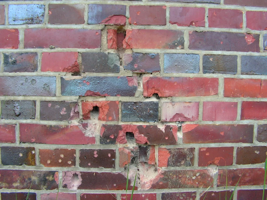 Ceglana ściana z licznymi ubytkami i uszkodzeniami, widoczne ubytki i zadrapania w cegłach oraz zaprawie wskazują na degradację materiału budowlanego.