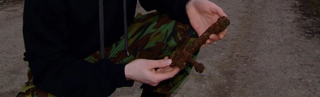 Osoba w wojskowych spodniach trzyma w dłoniach zardzewiały przedmiot przypominający fragment broni lub metalowy artefakt, tle widoczna ziemista droga.