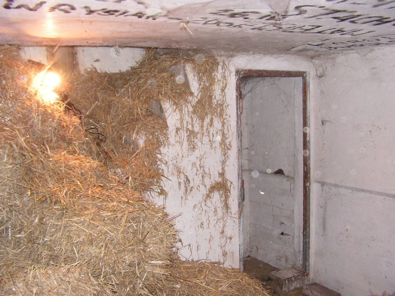 Stosy siana ułożone w białym, betonowym pomieszczeniu, słabo oświetlone żarówką wiszącą w rogu, otwarte drzwi prowadzą do kolejnego, równie zaniedbanego wnętrza.