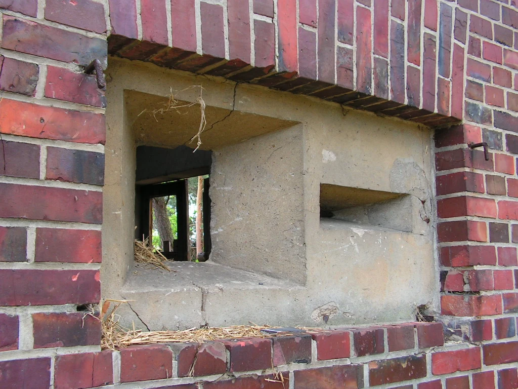 Otwór strzelniczy w betonowej ścianie obłożonej cegłą, widoczne wnętrze stanowiska obronnego z lekkimi śladami zużycia i resztkami suchej trawy lub słomy.