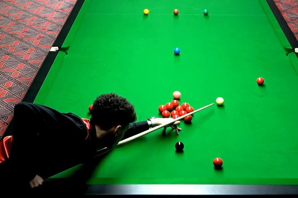 Mężczyzna skupiony na wykonaniu zagrania w snookera, trzyma kij bilardowy nad stołem wyłożonym zielonym suknem, otoczony kolorowymi bilami, na tle wzorzystej wykładziny.