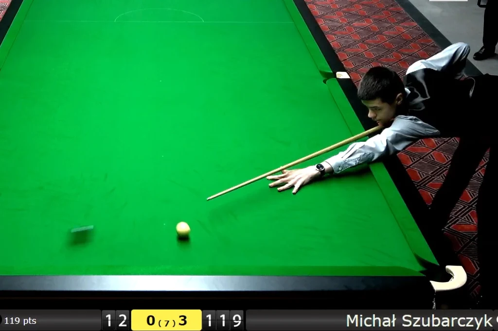 Gracz w trakcie uderzenia bilą snookerową przy zielonym stole, na ekranie widoczne są statystyki meczu i nazwisko zawodnika oraz punktacja.