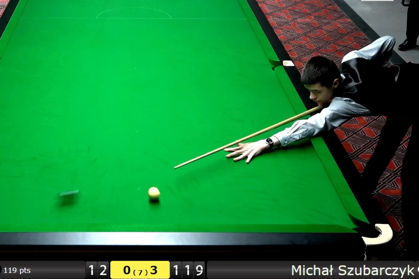 Gracz w trakcie uderzenia bilą snookerową przy zielonym stole, na ekranie widoczne są statystyki meczu i nazwisko zawodnika oraz punktacja.