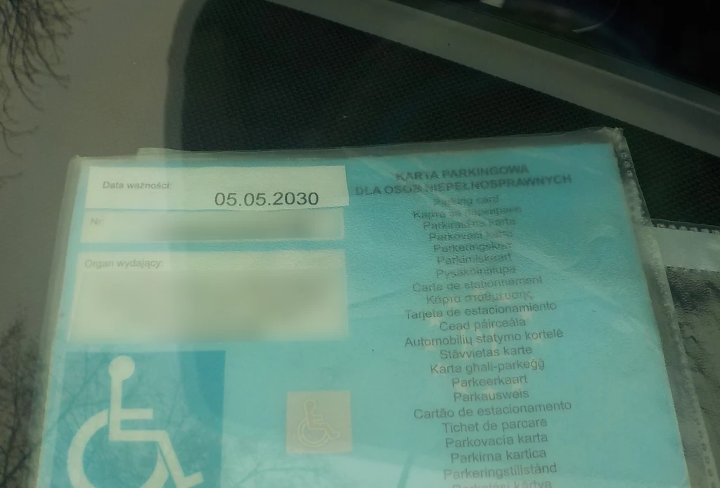 Niebieska karta parkingowa dla osób z niepełnosprawnościami z widocznym terminem ważności do 05.05.2030 oraz symbolem wózka inwalidzkiego, znajdująca się za szybą pojazdu.