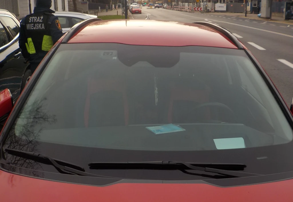 Zbliżenie na przednią szybę czerwonego samochodu, na której znajduje się niebieska kartka, w tle na chodniku funkcjonariusz straży miejskiej w mundurze, za samochodem widoczna jest ulica i budynki.
