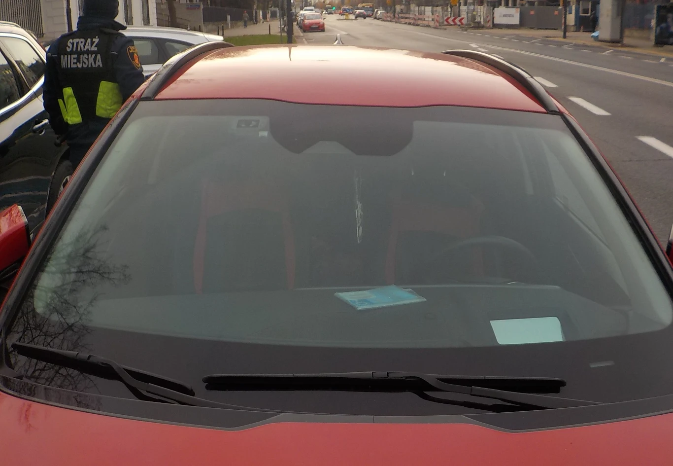 Na pierwszy rzut oka ciężko stwierdzić, czy karta jest podrobiona czy legalna / Fot: Straż Miejska w Warszawie Zbliżenie na przednią szybę czerwonego samochodu, na której znajduje się niebieska kartka, w tle na chodniku funkcjonariusz straży miejskiej w mundurze, za samochodem widoczna jest ulica i budynki.