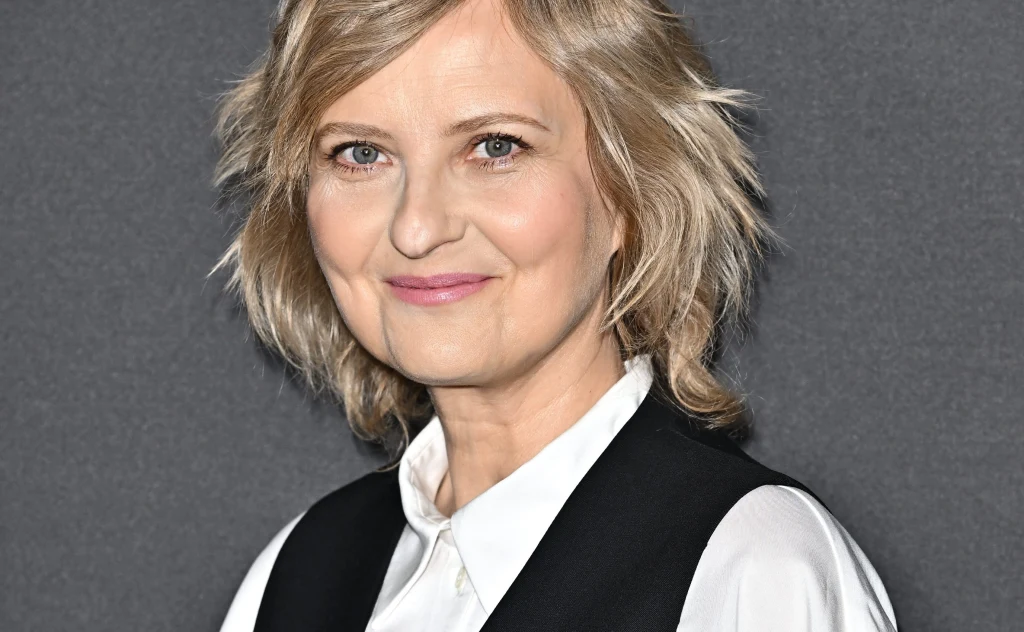 Uśmiechnięta kobieta z blond włosami o jasnych oczach, ubrana w białą koszulę i czarną kamizelkę, na tle ciemnej, jednolitej ściany.