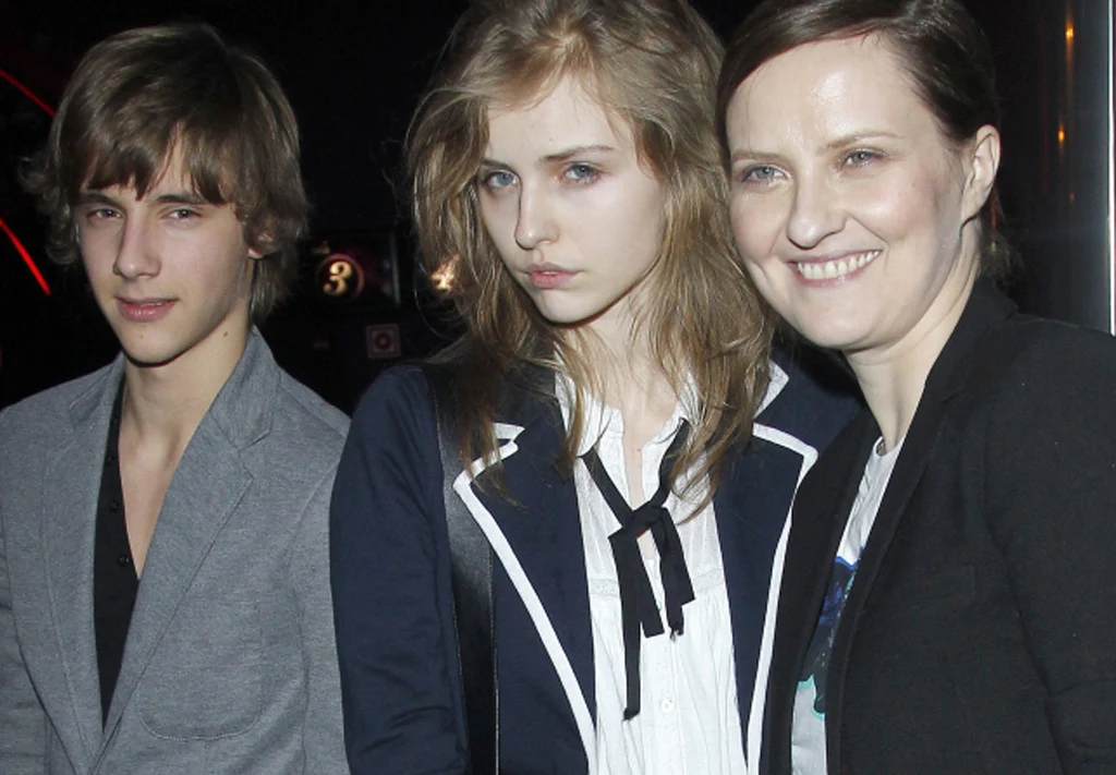 Trzy osoby stoją obok siebie, dwie z nich patrzą bezpośrednio w obiektyw, a jedna kobieta uśmiecha się szeroko. Osoby ubrane są elegancko, tło pozostaje ciemne i nieostre.