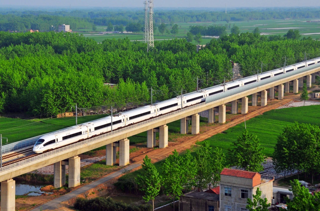 Nowoczesny biały pociąg dużych prędkości porusza się po długim wiadukcie nad zielonymi polami i gęsto zadrzewionym krajobrazem wiejskim, z pojedynczym domem w prawym dolnym rogu kadru.
