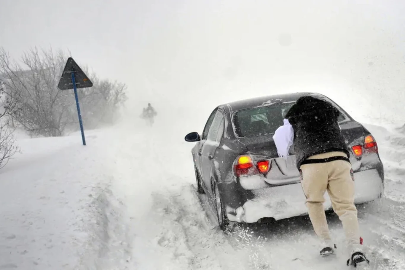 Samochód utknął na zaśnieżonej drodze, mężczyzna pcha auto przez intensywną zamieć, w tle niewyraźna postać oraz zasypane otoczenie, ograniczona widoczność przez śnieg i wiatr.