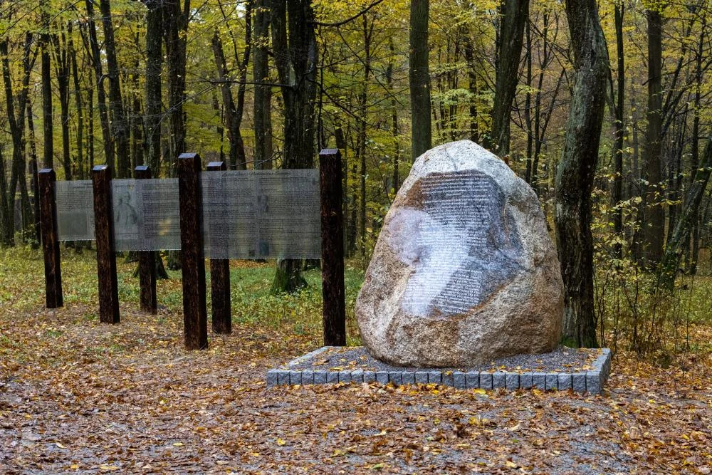 Nowy rezerwat przyrody Las Zapaśny upamiętnia leśników Duży głaz z wyrytymi napisami, stanowiący pomnik pamięci, stoi na brukowanym podeście w lesie, obok przezroczyste tablice informacyjne umocowane na drewnianych słupach. Otoczenie tworzy jesienny krajobraz z opadającymi liśćmi i drzewami.