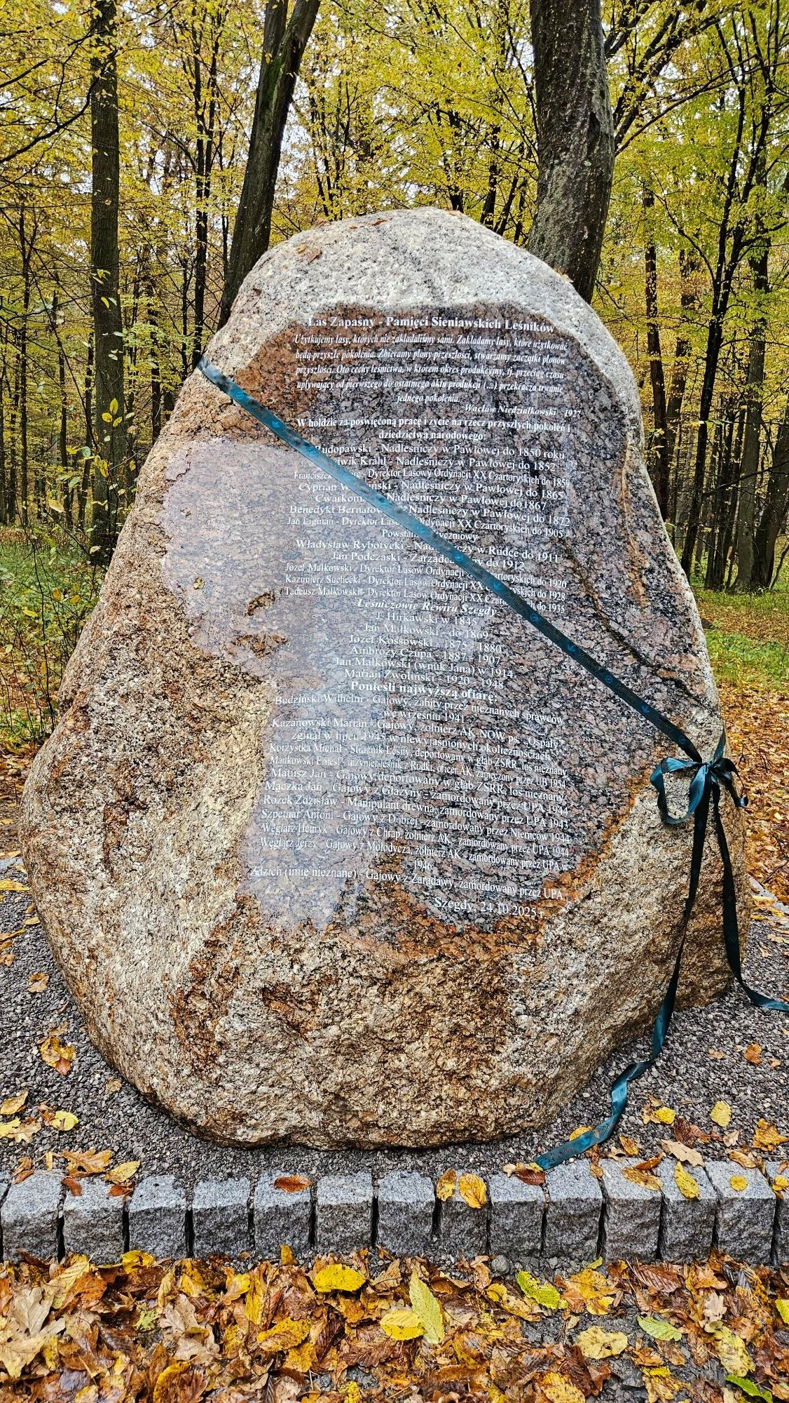 Kamień z inskrypcją Duży głaz pamiątkowy z wyrytą inskrypcją, przewiązany niebieską wstęgą, ustawiony na kamiennym podwyższeniu pośród jesiennego lasu.