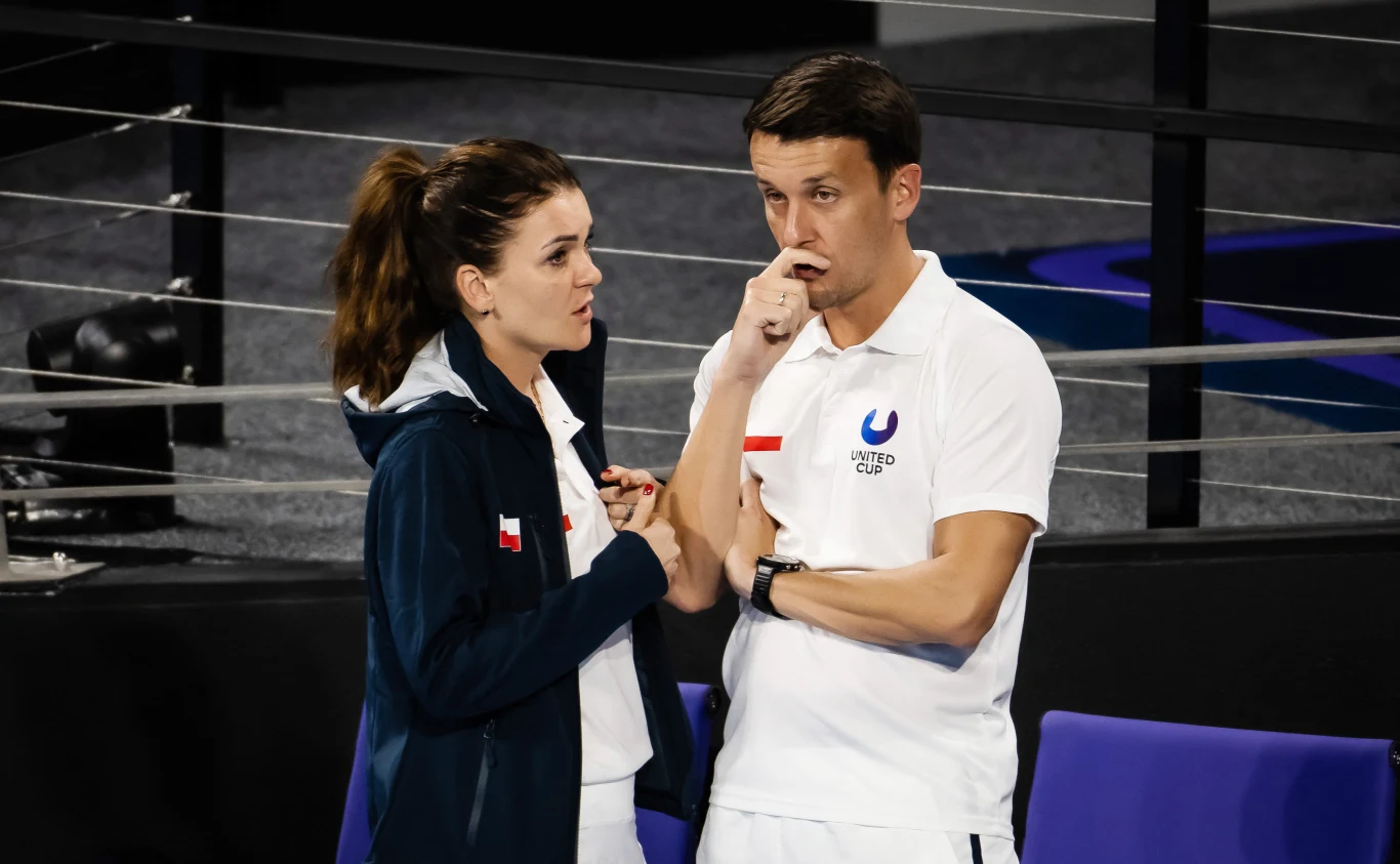 Dwie osoby w sportowych ubraniach prowadzą rozmowę w hali sportowej, kobieta w granatowej kurtce z polską flagą oraz mężczyzna w białej koszulce z logo i polską flagą, oboje skupieni na dyskusji.