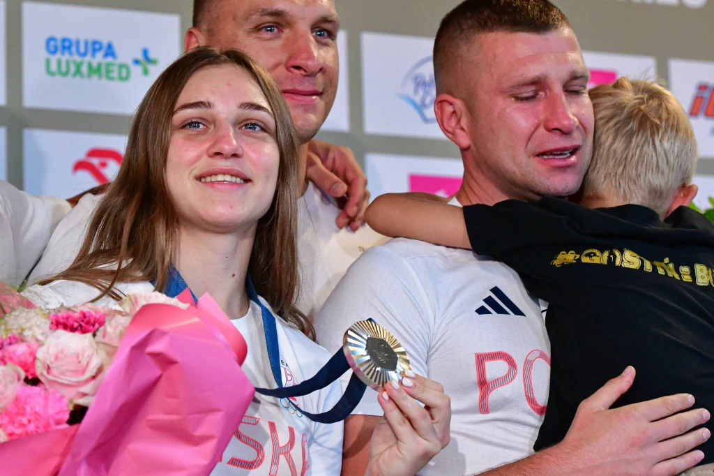 Julia Szeremeta i trenere Tomasz Dylak (z prawej) Srebrny medalistka z bukietem kwiatów uśmiecha się, obok jej wzruszony kolega przytula dziecko, w tle widoczne logo sponsorów.